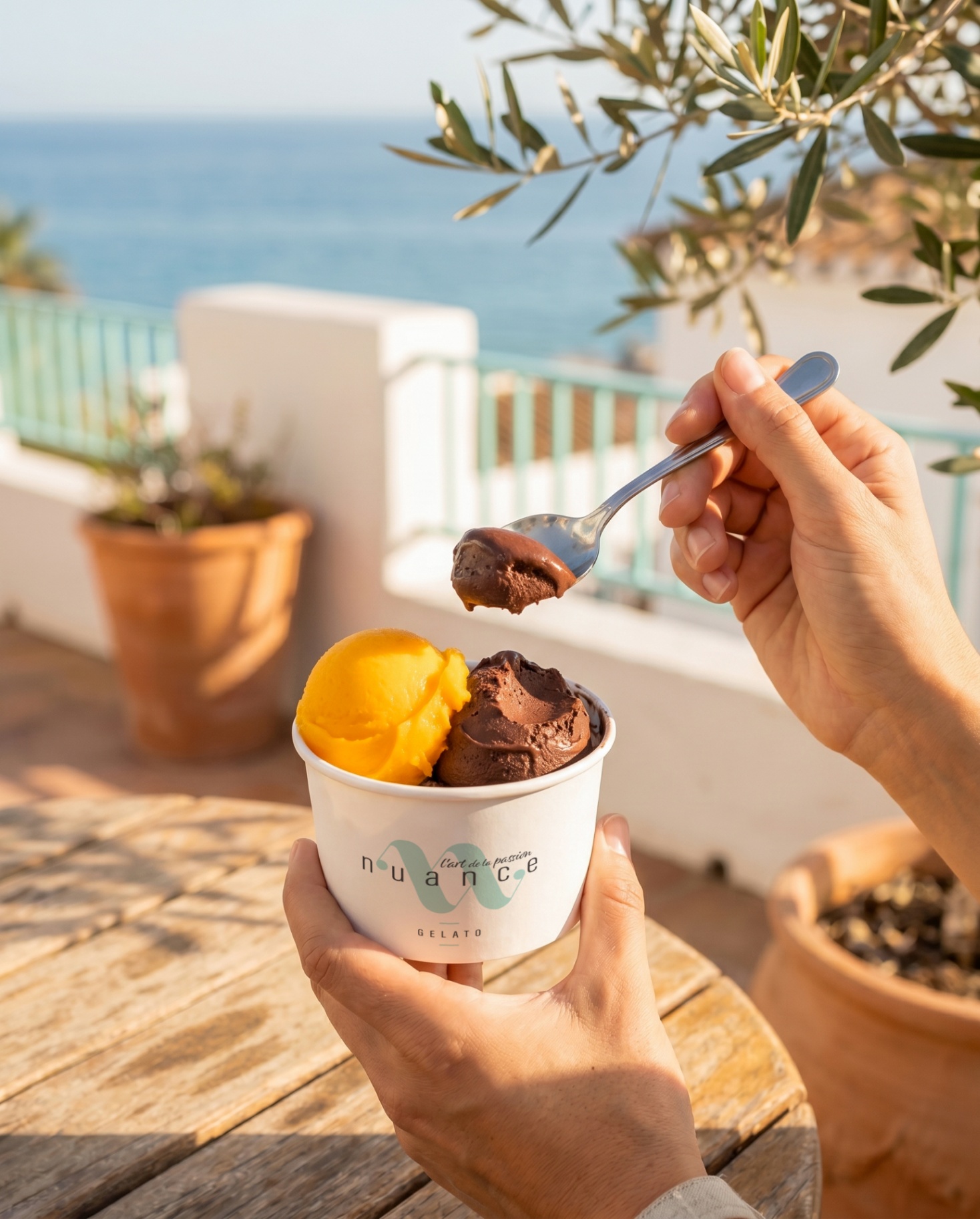 Hands holding a NUANCE cup with mango and chocolate gelato overlooking the sea